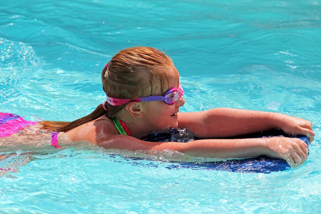 Großaufnahme eines Mädchens mit bunten Schwimmsachen und bunter Schwimmbrille schwimmt mit einem Schwimmbrett